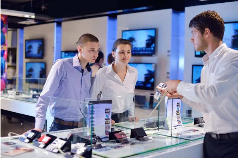 A sales representative demonstrating the features of a new smartphone to two customers in a high-tech electronics store, emphasizing the effectiveness of 3D product animation services in showcasing innovative device functions and design for marketing campaigns.